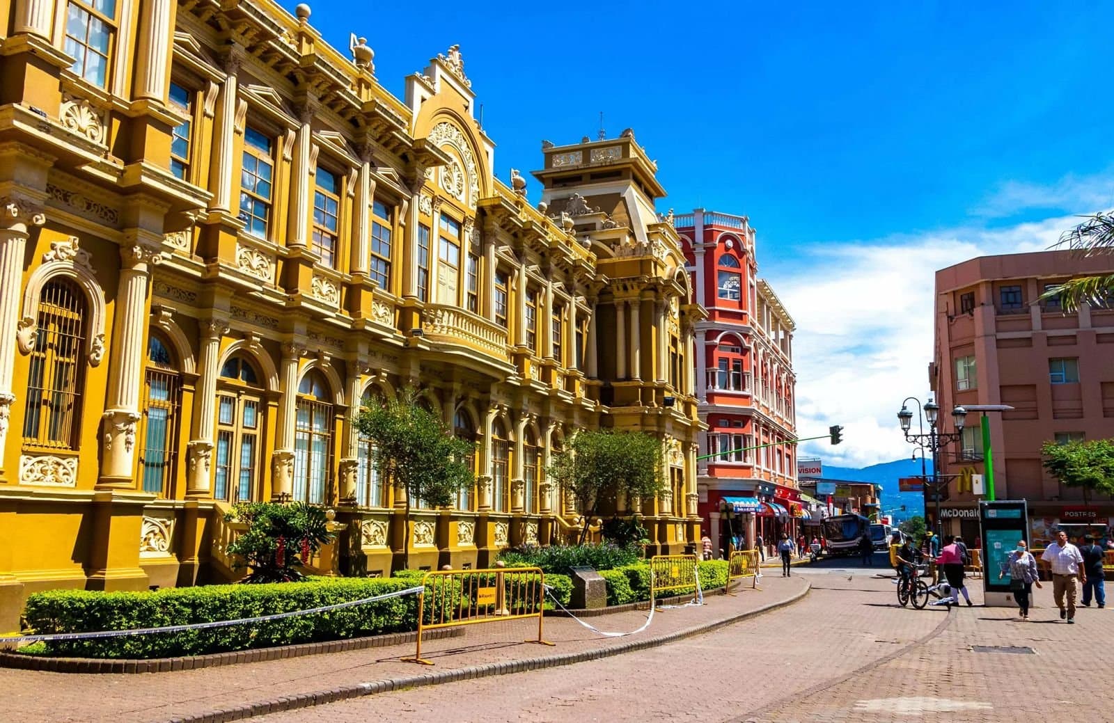 An ornate yellow colonial-era building on a street in downtown San José, with a red building and city traffic behind it